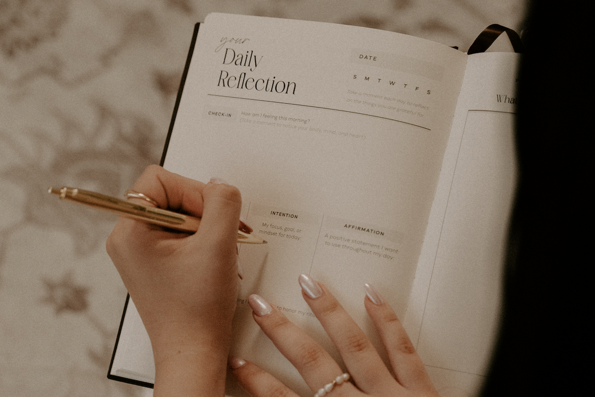 Person writing in a journal titled 'Daily Reflection' with a pen.